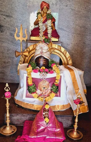 A shrine alcove with a golden statue of a seated figure in a red turban and red garments, one hand raised in blessing, adorned with white and green garlands. Below stands a dark stone Shiva linga draped in a white cloth with gold border, wearing a jewelled head ornament, with yellow marigold and green leaf garlands and pink flowers. A framed printed picture of a deity murti sits in front. A golden trident leans on the left, and brass oil lamps with pink flowers flank the base, with a pink silk cloth on the floor. No people are visible.