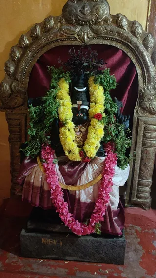 A dark stone murti of a Shiva form is decorated for worship inside an ornate brass arch set against a maroon velvet backdrop. The deity wears a crescent moon on its head, a vertical tilak mark, and a gold necklace. Garlands of yellow marigolds, pink flowers and green leaves drape across its shoulders and chest, with a sprig of dark leaves tucked on top. It is draped in a pink and cream silk cloth with maroon borders. The black stone pedestal is engraved with the letters NIEV-1, and the floor is red.