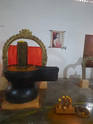 Interior shrine photo showing a black stone Shivalinga on a black yoni-peetha base, set within an ornate gold-coloured arched frame. The lingam wears a yellow flower garland and a dark rudraksha-style bead necklace, with a red cloth backdrop behind it. On the white wall at right hangs a small printed portrait of a smiling figure with long dark hair, wearing a pink-red garment and a white flower garland; the figure is a printed picture, not a physical person, and no name is legible on it. Small brass or bronze deity murtis with yellow flowers and a tray with a flower offering sit on the floor at lower right. No people are visible.