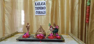 A photograph of a small decorated shrine set on a dark wooden platform before gold drapery. A sign reading KAILASA TRINIDAD & TOBAGO hangs on the drapery at top centre. On the left stands a small gold figure of a standing deity with a crown-like headdress, beside red and pink flower garlands. At centre is a gold stepped shrine form, a meru-like structure, surrounded by red and pink flowers. On the right are gold seated deity murti with a green cloth draped over the base, decorated with red and pink flowers and green leaves. A red flower garland hangs at the upper right edge. No people are visible.