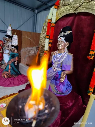 A photograph of a golden-toned deity murti seated on a crimson velvet throne with a gold ornate canopy decorated with orange and white flower garlands. The seated figure wears a purple and blue silk sari with a gold border, layered pearl and red-stone necklaces, earrings, a pearl-studded headdress with a small tilak, and a red bindi. In the foreground a lit oil lamp in a dark bowl burns with a tall flame. Behind on the left are two smaller metallic deity murtis wearing white and teal garments with tall decorated crowns. Yellow and red cloth covers the floor. No physical people are visible.
