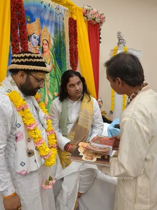 A photograph of three men in a decorated room with yellow and red drapes and a backdrop painting of Radha and Krishna with crowns and green garments. A man on the left wears a black and beige woven cap, white embroidered kurta with feather motifs, and a yellow and pink marigold garland, and looks down. A seated man in the centre wears a white kurta and dhoti with a yellow-and-beige shawl, has long swept-back dark hair and a red tilak, and holds a stack of books. A man on the right in a cream striped kurta wears a rudraksha mala and accepts the books. A silver deity murti sits on a pedestal behind them, with pink and white flower arrangements above.