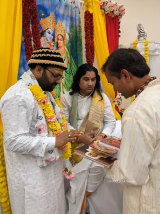 A photograph of three men in white traditional kurta clothing in a decorated room with yellow and red drapes and garlands of yellow marigolds and pink roses. Behind them a painted backdrop shows Krishna with a peacock-feather crown and Radha in green. The man on the left wears a striped black-and-cream cap, glasses, a white shawl with a garland of yellow and pink flowers, and a tilak on his forehead; he holds his hands together in a receiving gesture. The seated man in the centre wears a white outfit, a green and yellow shawl, long dark hair and a red bindi, and watches the hands. The man on the right, bending forward, wears a cream kurta and a rudraksha mala and holds a stack of books and booklets. A silver deity figure sits on a white pedestal at the upper right.