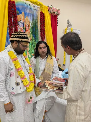 A photograph in a decorated room with yellow and red drapes, marigold and rose garlands, and a painted backdrop of Radha and Krishna with a peacock feather. A man with long dark hair and a red-orange tilak, wearing white kurta and pants with a green and gold shawl, sits centrally on a white cloth. On the left, a man in a white embroidered kurta, black-and-cream knitted cap and yellow-and-pink marigold garland stands looking down. On the right, a man in a cream striped kurta with rudraksha beads holds a stack of printed booklets with orange and red covers, offering them toward the seated man. A silver deity figure sits on a white pedestal at right.