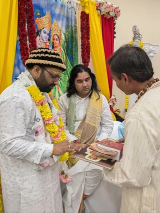 A photograph of a ceremony in front of a backdrop depicting Krishna playing a flute beside Radha, framed by red and yellow draped fabric and garlands of red roses and pink flowers. A man in a white embroidered kurta with a black-and-cream crocheted cap and a yellow-and-pink marigold garland stands at left, looking down and touching an open book. Seated centrally, a man with long dark hair, a red tilak on his forehead, a white kurta and a striped gold-and-brown shawl with a green wrap watches the proceedings. At right, a man in a cream kurta with a rudraksha mala holds up a stack of printed booklets. A small silver deity figure sits atop a white pedestal at the upper right. The third man is the one at right. Photo credit overlay reads KAILASA Seattle.