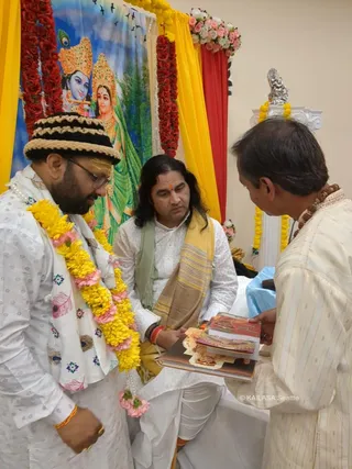 A photograph of three men in white ethnic clothing indoors before a painted backdrop of Radha and Krishna framed by red and yellow draped fabric and marigold garlands. On the left, a bearded man in a woven brown-and-black cap and a white kurta with peacock-feather print wears a yellow and pink flower garland and gold bangle, looking down. In the centre, a seated man with long dark hair, a white kurta, a green-and-gold striped shawl and a red tilak sits holding a stack of books and booklets. On the right, a man in a cream kurta with a rudraksha mala holds out the stack of printed material. A silver figurine sits on a white pedestal behind them with yellow flower garlands.