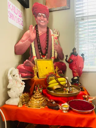 A home puja altar set against a beige wall, with an orange cloth draped over the table and a red satin runner beneath the offerings. A life-size cutout standing behind the altar shows a smiling man in a red turban, red robe, rudraksha mala and white flower garland, raising one hand and holding a trident; this is a printed depiction, not a person present. Before it sits a gold yantra plate, a white marble Ganesha seated on a lotus, a small gold meru-style shrine with a deity figure, brass bells and a bowl, a brass tray of bananas and rudraksha malas, and a tray of red powder. A black deity murti in a red polka-dot cloth stands at right beside a window with white blinds. A handwritten paper sign on the wall reads KAILASA SAN DIEGO.