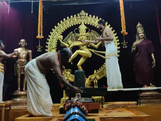 A photograph inside a temple shrine showing a golden Nataraja murti of dancing Shiva within an ornate golden arch, flanked by golden statues of a sage and a goddess in a maroon sari. Two bare-chested priests in white dhoti with red sacred threads perform abhishekam at a black lingam on a pedestal; the older priest in front bends to pour liquid over it while the younger priest at right stands on the raised platform beside the Nataraja. Marigold garlands hang at the top corners, small oil lamps dangle at the sides, and red flower petals and floral offerings lie around the base. A watermarked logo sits at the lower right of the image.