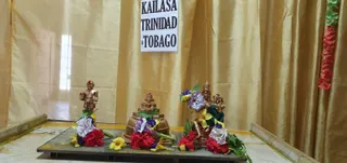 A photograph of a shrine arrangement in front of gold draped curtains. A paper sign pinned to the curtain reads KAILASA TRINIDAD & TOBAGO. On a dark wooden tray stand three gold-toned deity murtis: a small standing figure on the left, a tiered stepped structure in the centre, and a seated deity with a crown and green cloth on the right. Each group is decorated with red, yellow and purple flowers, white paper blossoms and green leaves. Red flower garlands hang on the right edge. No people are visible.