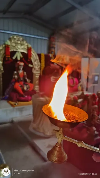 A brass oil lamp with a tall flame and rising smoke is held in the foreground of a temple shrine. Behind it, a seated deity murti in an orange and red garment sits beneath a gold arched canopy draped with red and orange garlands. Other red-draped altar items and offerings are visible in the background. No people are clearly visible; only a hand at the right edge holds the lamp. The photo appears to be an aarti at a shrine.