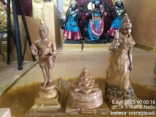 Photograph of a temple interior showing three bronze-toned statues on a golden-brown floor in front of a shrine. On the left stands a male statue with raised right hand in abhaya mudra and a dhoti-like lower garment. On the right stands a female statue draped in a flowing garment and holding a long staff. At the centre is a tiered stepped pyramid base, a shri yantra-like form of a sri chakra or meru shape on a round platform. Behind them, three dark-complexioned deity murtis in blue and pink skirts stand in an alcove, one holding a staff. Metal stands and golden decor are at the edges. Timestamp in the lower right reads 5 Apr 2025 10:00:16, with Tamil Nadu and kailasa seeragapadi labels.