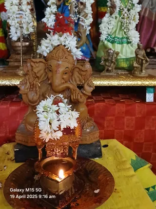 A photograph of a terracotta-coloured Ganesha murti seated on a red checked cloth before a lit oil lamp in a brass dish. The murti wears a white jasmine garland and a red flower garland, and the lamp flame is lit at its base on a yellow cloth. Behind it stand a garlanded figure in green and blue clothing, a brass kalasha with a garland, and small metal figurines on a gold-coloured altar. A date stamp reads 19 April 2025 9:16 am. No people are visible.