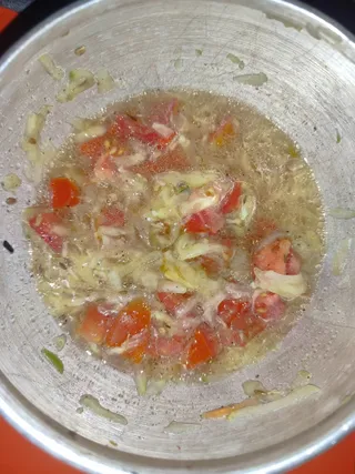 Close overhead photo of a stainless steel pot holding a bubbling, oily liquid cooking mixture. Diced red tomatoes and shredded pale vegetable. Bubbles cover the surface. A red cooktop surrounds the pot. No people, no swami or ritual setting visible; the image shows a cooking pot only.