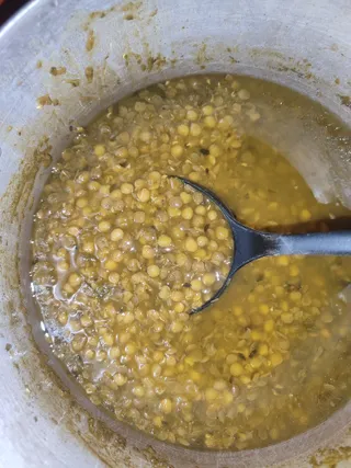 Close-up photo of a large steel cooking pot holding yellow split lentil dal, with whole and split yellow pulses floating in a golden broth. A black plastic ladle rests in the pot. No people are visible; the frame shows only food and cookware, with specks of spice and residue on the pot's inner rim.