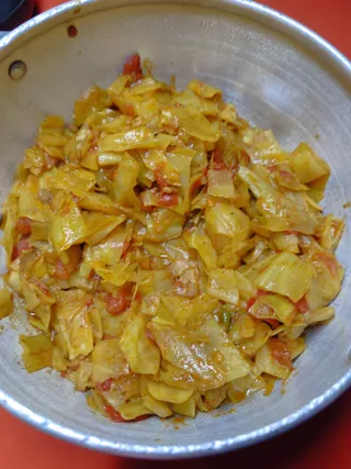 A close overhead photograph of a cooked vegetable dish in a large silver aluminium pot or pan, set on a bright red surface. The dish is shredded cabbage cooked with diced tomato pieces and turmeric-yellow seasoning, with glossy oil and spices coating the pale yellow-green cabbage strips. No people, a person, ritual setting or readable text is visible.