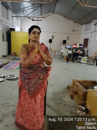 A photograph of a woman standing in a large, plain hall with a corrugated metal roof, pressing her palms together in an anjali greeting. She wears a red and maroon floral saree with a patterned border and a brown embroidered blouse, with gold bangles and a gold necklace, and has a white vibhuti marking with a red bindi on her forehead. Behind her are a yellow panel, a small table with a monitor and speakers, chairs, and a framed picture on the wall. At right is a brick-lined fire pit with a metal bowl, and plates with offerings lie on the floor at left.