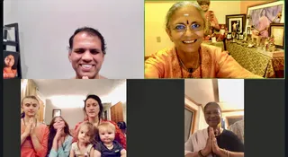 A multi-panel video call screenshot of a virtual yoga and puja session. Top-left panel shows a smiling man with short dark hair against a plain pale wall, a framed photo to his left. Top-right panel shows a smiling woman with grey hair, white vibhuti tilak on her forehead, glasses, an orange floral kurta and a beaded mala, seated before an altar with framed portraits, a deity murti, brass lamps and a gold-patterned cloth. Bottom-left panel shows four people with palms pressed in anjali: a young woman in orange with a tilak, a girl in a teal top, a woman in red with a tilak, and two small children. Bottom-right panel shows an older woman in a white shawl and black top, smiling with palms joined. Several people are visible across the panels.
