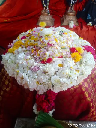 A large round arrangement of white, yellow, pink and red flowers heaped on a red cloth with gold dots, with a long red and pink flower garland hanging from its base onto a green leaf. Behind it, two copper-toned deity murti stand on pedestals before red drapes, one wearing a beaded necklace with white and green beads. A date stamp in the corner reads 12/13/25 07:58. No people are visible.