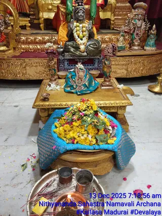 A photograph of a shrine interior with a dark metal murti seated on a pedestal, hands raised in blessing, wearing a garland of white flowers and a necklace with large yellow beads. Its body is draped in a green cloth. In front, a gold low table holds a blue silk-draped ornamented necklace, a small bell-metal bowl, and a heap of yellow marigolds and red petals sits on a blue brocade cloth. Two small brass attendant figures in red headdresses flank the table. In the foreground a brass plate holds copper vessels, incense sticks and a lit oil lamp. Gilded decorative stands and colourful statues stand behind. Petals are scattered on the floor. A digital timestamp and caption overlay the lower right corner.