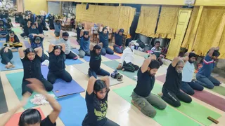A photograph of a yoga session in an indoor hall with pale wooden flooring and yellow-gold draped fabric on the walls. Participants kneel on colourful yoga mats in rows, arms raised overhead with hands clasped behind the head in a stretch. Visible clothing includes black t-shirts and black trousers, a girl in a black graphic tee, a man in a light blue shirt, and a man in a white shirt and dark trousers wearing a face mask. Some people are seated at the back near chairs and bags, and a few partially visible figures stand or sit along the edges. Mats are blue, purple, green, grey and red.