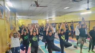 A photograph of a large yoga class in a yellow-walled hall with ceiling fans and fluorescent lights. Many participants stand on colourful rolled mats in tree pose with palms pressed together overhead. Most wear black t-shirts, one with a green graphic reading Moment Enjoy, another in a white shirt and dark trousers, and a man on the right in a black shirt and olive trousers balances on one leg. Framed pictures hang on the left wall. No identifiable person is shown as a person in this image.