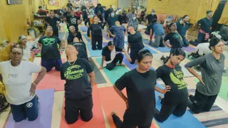 A photograph of a yoga session in a large hall with yellow walls and a golden curtain at the top. Many participants, mostly women and some men, kneel and stretch on colourful rectangular mats in red, green, blue, purple and grey. Foreground: a woman in a white t-shirt with a tilak on her forehead; women in black t-shirts, one reading 'Every Moment Enjoy' in yellow letters and another with a yellow eagle graphic; a woman in a grey top; a woman in a white sari-like wrap kneeling. Several participants wear face masks. Background shows people seated along walls and a small shrine area with framed images.