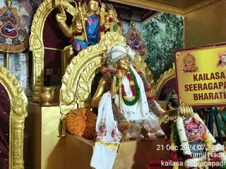 A golden statue of a seated sage in a white turban and white robe with a garland of red, white and green flowers sits on a gilded throne in a temple shrine. The figure holds a golden staff and raises one hand in blessing. Behind him a gilded deity murti with a trident is visible, and a small oil lamp burns on the ledge. To the right stands a yellow board reading KAILASA SEERAGAPADI and BHARATH, beside a framed photo of a saffron-robed man. Marigold garlands and a red-and-green floral arrangement decorate the scene. The timestamp shows 21 Dec 2024 07:32:34, Tamil Nadu.