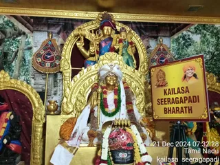 A photograph of a shrine with gilded statues, no living people visible. A golden statue of a seated figure in a white turban, white dhoti-style cloth and a green and red-white garland holds a trident-like staff and sits before a black lingam draped in a red cloth and white and yellow flower garlands, with a brass kalasha beside it. Above, a gilded deity murti with a trident and a consort figure in a green sari is set in an ornate golden arch. To the right, a yellow board reads KAILASA SEERAGAPADI BHARATH beside a small portrait of a smiling man in an orange turban and orange clothing. Painted forest backdrops and gold-framed panels surround the shrine.