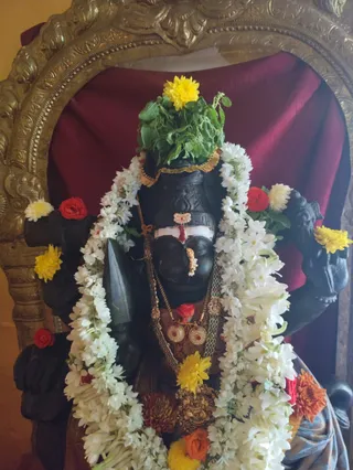 A dark stone deity murti is decorated for worship, framed by an ornate gold arched niche against a deep maroon cloth backdrop. The murti wears a long white jasmine garland, a crown-like headpiece of green leaves topped with a yellow marigold, and gold ornaments including a nose ring, pendants and a jewelled necklace. Red roses and yellow and orange marigolds are placed around the figure, with a red tilak on the forehead. No people are visible.