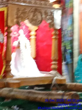 Blurred photo of a white-clad figure seated on a wooden platform before a red backdrop with carved wooden pillars and pink floral garlands. The figure wears white garments and a white and pink garland, appearing to be a deity murti or decorated statue rather than a living person. Appearance cues are insufficient to identify a person candidate.