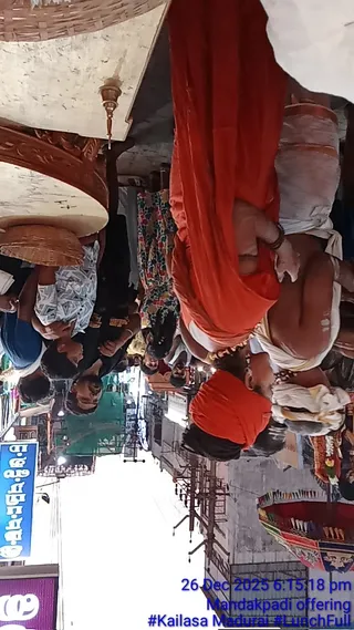 A photograph taken in an outdoor street procession, rotated sideways in the frame. A man in an orange-red shawl and white dhoti stands at the centre-right, bare-chested with sacred markings on his body and beaded necklaces, while a second figure in an orange turban and dark clothing is seen below him. Several people in light-coloured shirts and dark clothing stand at left beneath a large wicker basket-like canopy and an ornate wooden structure with a brass lamp hanging down. A decorated cloth-draped vehicle with coloured banners appears at right, and a tall overhead structure with electric poles lines the street. Bright sunlight lights the scene, and a blue signboard with non-Latin script sits at left. Blue timestamp text at lower right reads 26 Dec 2025 6:15:18 pm and Mandakpadi offering.