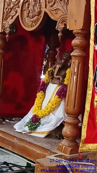 A photograph of a carved wooden palanquin or shrine canopy with ornate carved pillars and top panels, framing a red cloth backdrop. Inside sits a dark wooden murti draped in a white cloth with a gold border, decorated with a yellow marigold garland and a purple flower garland, with green leaves tied at its base. A red and gold embroidered cloth hangs on the right side. The bottom-right corner carries a blue timestamp reading 27 Dec 2025 6:05:38 pm and Mandakpadi offering. No physical people are clearly visible in the frame.