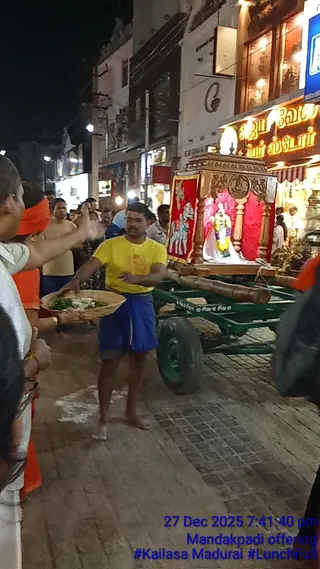 Night street procession in Madurai. A man in a yellow t-shirt and blue shorts walks barefoot on paving stones, holding a woven basket of food and gesturing with his right hand. Behind him a green wheeled cart carries a wooden canopied shrine with carved panels and red drapery, containing a white deity murti garlanded with yellow flowers. To the left a person wears an orange head cloth and white clothing, and other onlookers stand along the street under shop lights. Illuminated shop signs with Tamil lettering line the street.