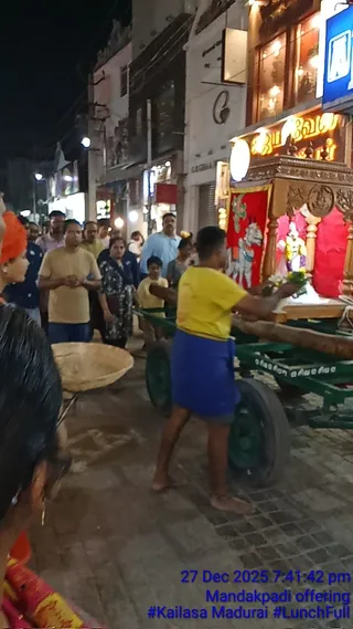 Night street photograph of a Mandakpadi procession in Madurai. A young man in a yellow t-shirt and blue cloth wrapped at his waist pushes a green wooden cart or palanquin decorated with a red cloth and a small deity murti adorned with flowers. A shaved-headed man in an orange head covering stands at the left edge, and a crowd of onlookers in casual clothes, including a woman in a dark patterned sari and a child in yellow, walks along the street lined with shopfronts and lights. A large woven basket sits on the left. Overlay text reads 27 Dec 2025 7:41:42 pm, Mandakpadi offering, #Kailasa Madurai, #LunchFull.