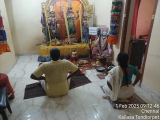 Indoor photo of a small shrine room with white marble floor and cream walls. Two devotees sit cross-legged on woven mats facing a shrine: a man in a yellow shirt and white dhoti at left, and a woman in a teal blouse and white sari with a long braid at right. The shrine holds three painted deity murtis adorned with flower garlands, on a gold-draped platform with brass oil lamps, a kalasha, plates of offerings and a small kumkum/vibhuti setup. To the right stands a printed portrait of a bearded sadhu with long hair and a rudraksha mala, beside a white sign reading KAILASA TONDIORPET. A pale orange cloth hangs at the right edge. A timestamp reads 1 Feb 2025 09:12:46 Chennai, Kailasa Tondiorpet.