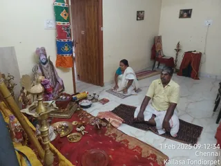 A photo of a small home shrine room with cream walls and a white marble floor. On the left, a standing figure in pink and orange ceremonial clothing, with a pink turban, rudraksha-style beads and a flower garland, stands beside a brass lamp and a red-draped altar laden with brass plates, flowers, offerings and a kalasha-like pot. A woman in a white saree with a gold border sits cross-legged on a mat near the wall. In the foreground, a man in a yellow shirt and white dhoti sits in meditation on a dark patterned mat, hands resting on his knees. Framed pictures hang on the walls and a brass lamp stands on a stool by a red cloth on the right. A timestamp reads 1 Feb 2025 09:12:34 Chennai Kailasa Tondiorpet.