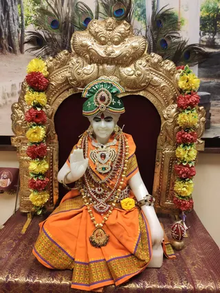 A white marble murti seated in a gilded arched shrine framed with red and yellow flower garlands and peacock feathers above. The figure wears a green velvet turban with jewelled ornaments, an orange silk dhoti with gold border, layered gold and jewelled necklaces and bangles, and raises one hand in abhaya mudra. A brass kalasha stands at the right and a patterned maroon cloth covers the base. The image is a deity murti, not a living person, and no one else is present.