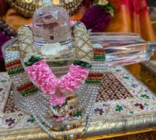 Close photograph of a clear crystal Shivalinga on a decorated crystal base, dressed with a pink satin rose garland with green leaves, a white rose, and strands of pearls, red and green beads, and gold-toned ornamental clasps. The linga has a small red-brown mark and a gold-trimmed ornament, and a white flower rests at its base. Behind it sit a red-and-gold fabric and blurred purple flowers. No people are visible; the scene is a ritual adornment of a sacred linga.