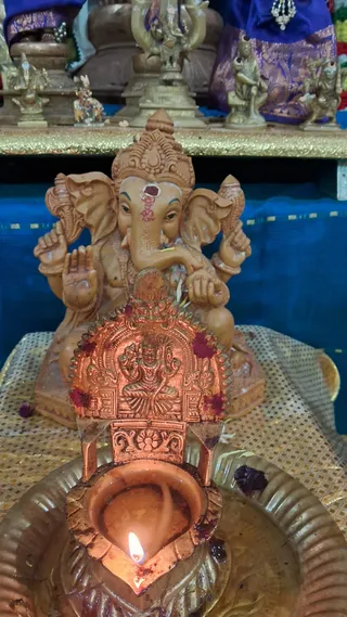 Close photo of a small terracotta-coloured Ganesha murti seated on a pedestal, with a red tilak on its forehead, a crown, and raised hands holding objects. In front, a copper-toned oil lamp with a lit flame and a decorated copper frame sits on a brass plate. Behind, a blue cloth backdrop, brass figurines and a purple silk textile are visible on a shelf. No people are visible.