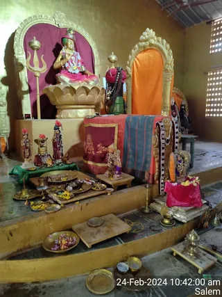 Interior photo of a shrine room with a golden arched niche and a golden lotus pedestal holding a seated murti dressed in pink and garlanded with white flowers, its hands raised in blessing; a golden trident stands beside it. To the right, a second golden-arched niche with orange cloth frames another murti in red and green attire. In the foreground, brass plates hold marigold and flower petals, brass bells and oil lamps sit on a wooden platform, and a pink velvet cloth covers a small altar. Timestamp reads 24-Feb-2024 10:42:00 am Salem. No people are visible.