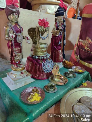 Photograph of a ritual display on a green brocade cloth at a KAILASA temple. Two gilded standing figures in maroon silk with gold-brocade borders and silver jewellery flank a central gilded cobra-hooded form with red hibiscus flowers, all draped in maroon silk and silver ornaments. In front are brass oil lamps with lit wicks, small brass bowls, a copper plate with a gilded figure and yellow flowers, and a brass tray. A maroon cloth with a printed bull motif hangs at the right. No people are visible; the figures are statues. Timestamp reads 24-Feb-2024 10:41:13 am, Salem.