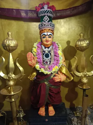 A standing painted clay or stucco murti of a deity in a temple shrine, set against a golden wall with a maroon cloth canopy above. The figure wears a tall green and silver crown decorated with red flowers, a pink and yellow marigold garland, a layered silver and gold necklace with a dark central pendant, maroon dhoti, and gold armbands. The right hand is raised in abhaya mudra with red painted markings, the left hand rests at the waist. Gold trident-topped lamp stands flank the figure, with a small brass oil lamp at the base. No people are visible.