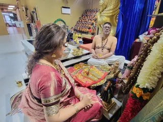 A photo inside a temple-like room with a golden statue of a mythic beast and a shelf of small brass deity figures behind it. In the foreground a woman in a maroon and gold silk sari with gold bangles and earrings sits with her hands clasped, facing a brass puja tray that holds an ornate padukas-style wooden sandal pair, marigold petals, a small lamp and kumkum. Beside her, a bare-chested man with a grey beard and glasses, wearing white dhoti cloth and several beaded malas, sits cross-legged facing the tray; his appearance does not clearly match the a person cues, so he is described only as a priest or devotee. Garlands of white flowers and orange marigolds hang on the right beside a blue curtain, and a brass kalasha vessel and a murti of a seated deity sit on the altar.