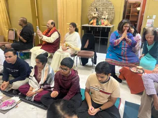 Indoor photo of devotees seated during a puja at KAILASA Toronto. In the foreground, a man in a navy Tommy Hilfiger sweatshirt and a young man in a cream Tommy Hilfiger t-shirt sit cross-legged on mats, one with eyes closed. A young woman in maroon with a white shawl and a teenage boy in a maroon hoodie and glasses sit nearby. Behind them, an older man in a maroon vest with orange shawl holds up a phone, beside a woman in a white sari. A woman in a blue patterned kurta sits with palms pressed together in namaste, next to a woman in teal. On the right, a red-cloth table holds a plate of offerings. Behind it, a small altar displays two painted deity murti figures under a round gold backdrop. Gold walls and cushioned chairs fill the room.