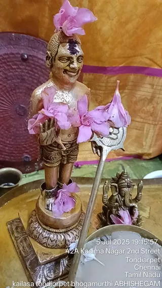 A small brass or bronze standing figure of a male deity murti, bare-chested with a dhoti-style lower garment, a tilak mark on the forehead and pink flowers draped across its shoulders, hands and feet, placed on a pedestal on a brass tray. A trident-like staff rests against it. Beside it sits a small Ganesha murti with pink flowers, and a metal bowl holds white liquid. The background is a saffron and magenta cloth. A timestamp and address overlay reads KAILASA TONDIARPET Bhogamurthi ABHISHEGAM.