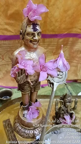 A small gilded metal murti of a standing figure with a vertical tilak on the forehead, adorned with pink flowers on the head, chest, arms and base. The figure holds a silver trident (trishul) in one hand and an open palm in the other. Beside it sits a small brass Ganesha figure with pink flowers, on a brass tray with a gold textile backdrop and a purple border. A timestamp overlay reads 5 Jul 2025 19:16:06 and the location Tondiarpet, Chennai. This is a photograph of a deity murti being bathed and decorated, not of a living person.