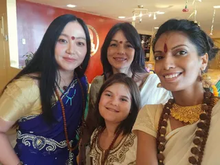 Four women and a young girl pose close together for a selfie in an indoor room with a red wall and a partly visible circular logo reading KAILASA. The woman on the left wears a royal blue sari with silver embroidery, a gold-brocade blouse, a turquoise bead necklace and a rudraksha mala, with a red bindi. Behind her a woman in a white and gold sari wears a red bindi. On the right a smiling woman in a cream sari with gold brocade wears a gold choker necklace, a long rudraksha mala and a red tilak on her forehead. A young girl in the centre wears a cream embroidered outfit with a rudraksha mala. Hanging floral decorations appear at the upper right.