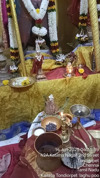 A photograph of a shrine altar decorated for daily worship. A small metal deity murti in a red garment and ornate crown stands at centre on a gold brocade cloth, flanked by white and yellow flower garlands hung on gold pillars. Brass plates hold marigold and white flowers, and a small lamp burns beside the murti. In the foreground, a brass lamp stand with a lit flame, a brass bowl of dark dried fruit or seeds, a white-filled bowl, a copper vessel, a coconut decorated with flowers and leaves, and a brass kalasha-like vessel are arranged on red cloth. A timestamp and location overlay appear at lower right. No people are visible.