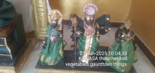 Photograph of three black stone deity murtis on a dark pedestal inside a temple shrine. The central deity wears a tall crown with white and pink beadwork, a gold and red chest ornament, and a green silk dhoti with gold border, holding a spear-like staff. The two flanking figures wear green silk garments with gold zari borders, jeweled crowns and pink flower garlands. A gold-toned wall and carved panel frame the shrine. A timestamp and handwritten-style caption overlay the lower centre. No people are visible; the image shows statues only.
