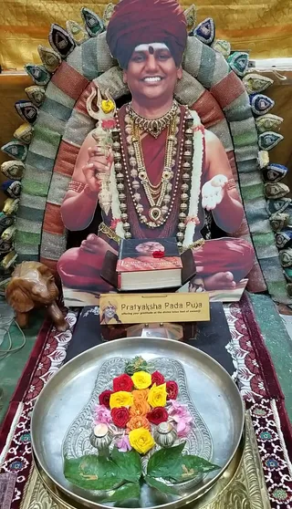 A photograph of a life-size cutout poster of a smiling man seated in a cross-legged pose against a tiered, padded backrest of striped and jewel-toned fabric. The figure wears a deep red turban, a red robe, layered gold necklaces, and strings of silver beaded malas, and holds a trident decorated with a yellow rose in one hand. A red-covered book titled Living Enlightenment sits in front of him on a wooden stand, with a red rose on top. Below is a sign reading Pratyaksha Pada Puja, offering your gratitude at the divine lotus feet of swamiji. In the foreground a round silver tray holds a floral arrangement of red, yellow and orange roses, pink blossoms, betel-type leaves and two small silver containers, on a red-bordered patterned cloth. A carved wooden elephant stands at the left edge. No physical people are present.